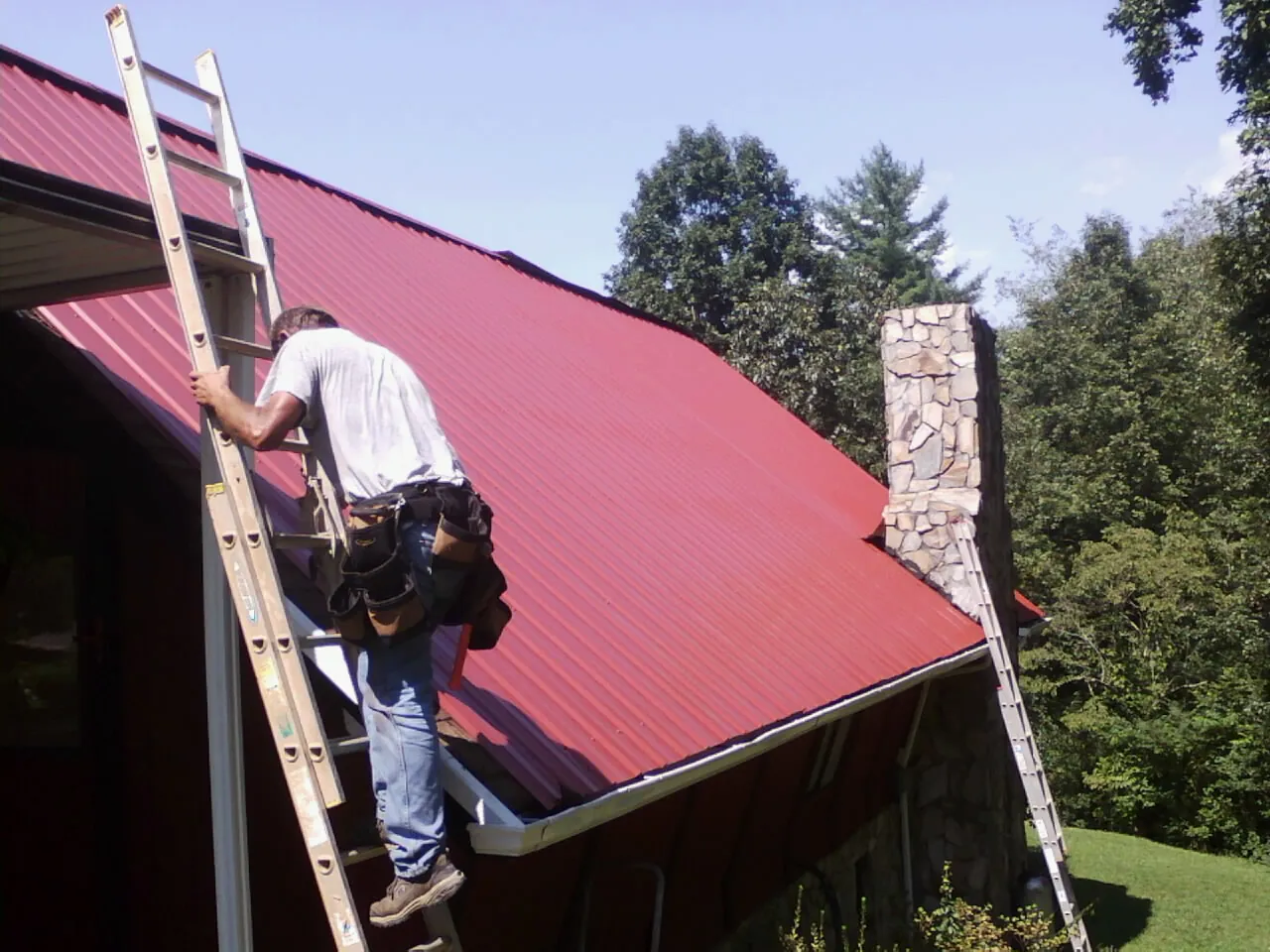 Roofer on a ladder beside a red metal roof and stone chimney — Soffit & Fascia Repair by Roofing Brothers in North Grosvenordale